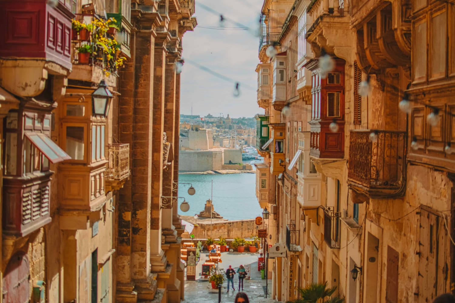 Narrow Valletta street flanked by limestone buildings and ornate wooden balconies, looking down toward the Grand Harbour